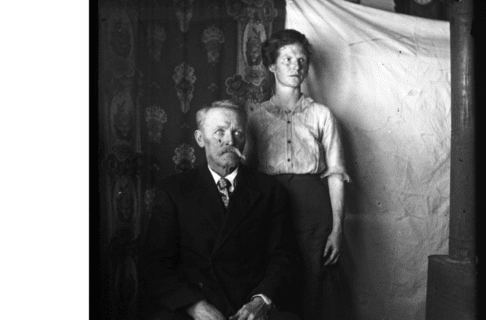 Black and white vintage photograph of a couple in front of a makeshift hanging backdrop. The moustachioed man is sitting, wearing a dark suit and tie. At his shoulder stands a woman wearing a button-up blouse and long skirt. Both have serious expressions and are looking slightly out of frame to their left.