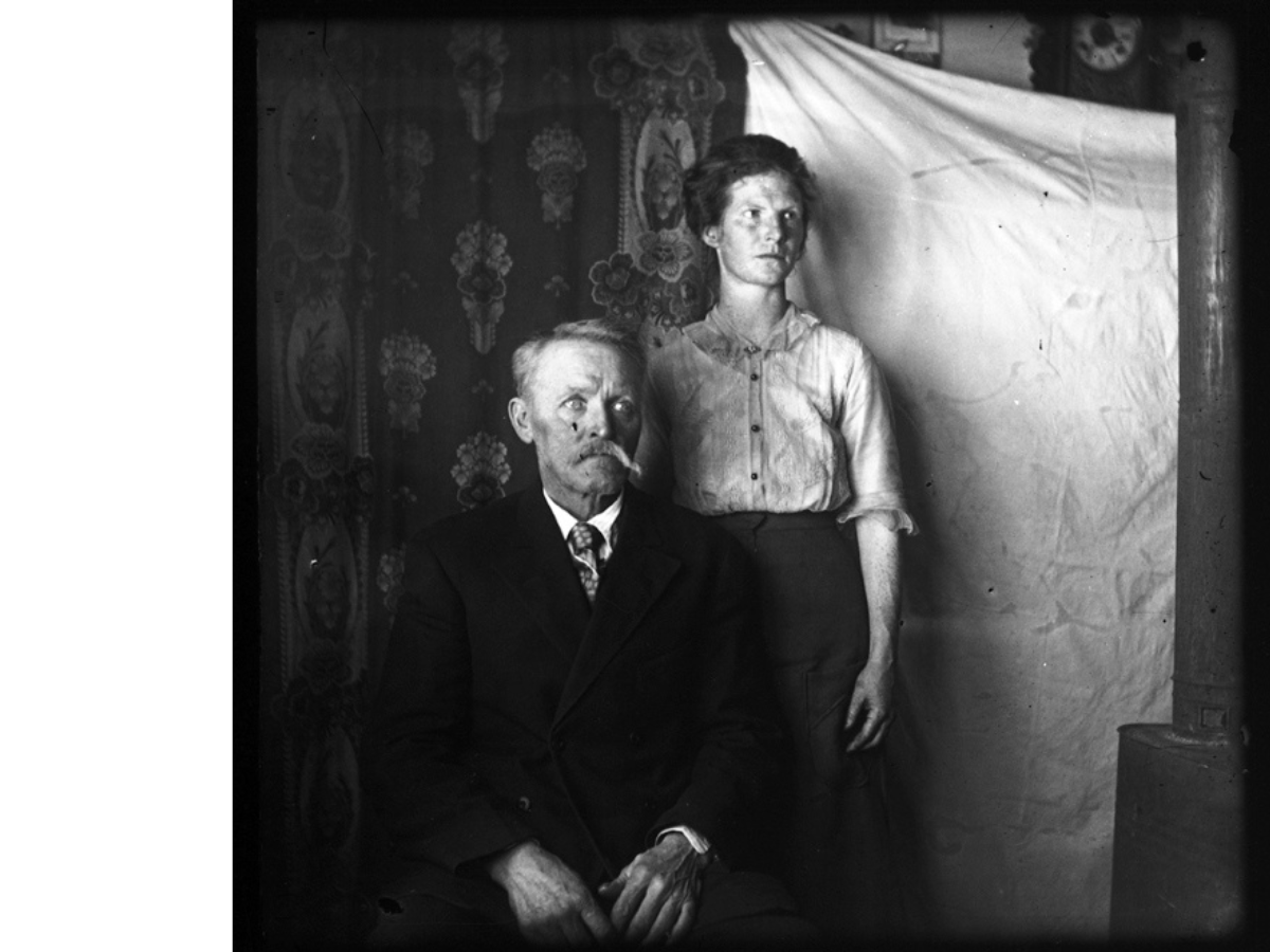 Black and white vintage photograph of a couple in front of a makeshift hanging backdrop. The moustachioed man is sitting, wearing a dark suit and tie. At his shoulder stands a woman wearing a button-up blouse and long skirt. Both have serious expressions and are looking slightly out of frame to their left.