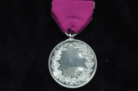 Photograph of a circular silver medal an oak leaf wreath. Medal is hung on a piece of purplish fabric or ribbon.