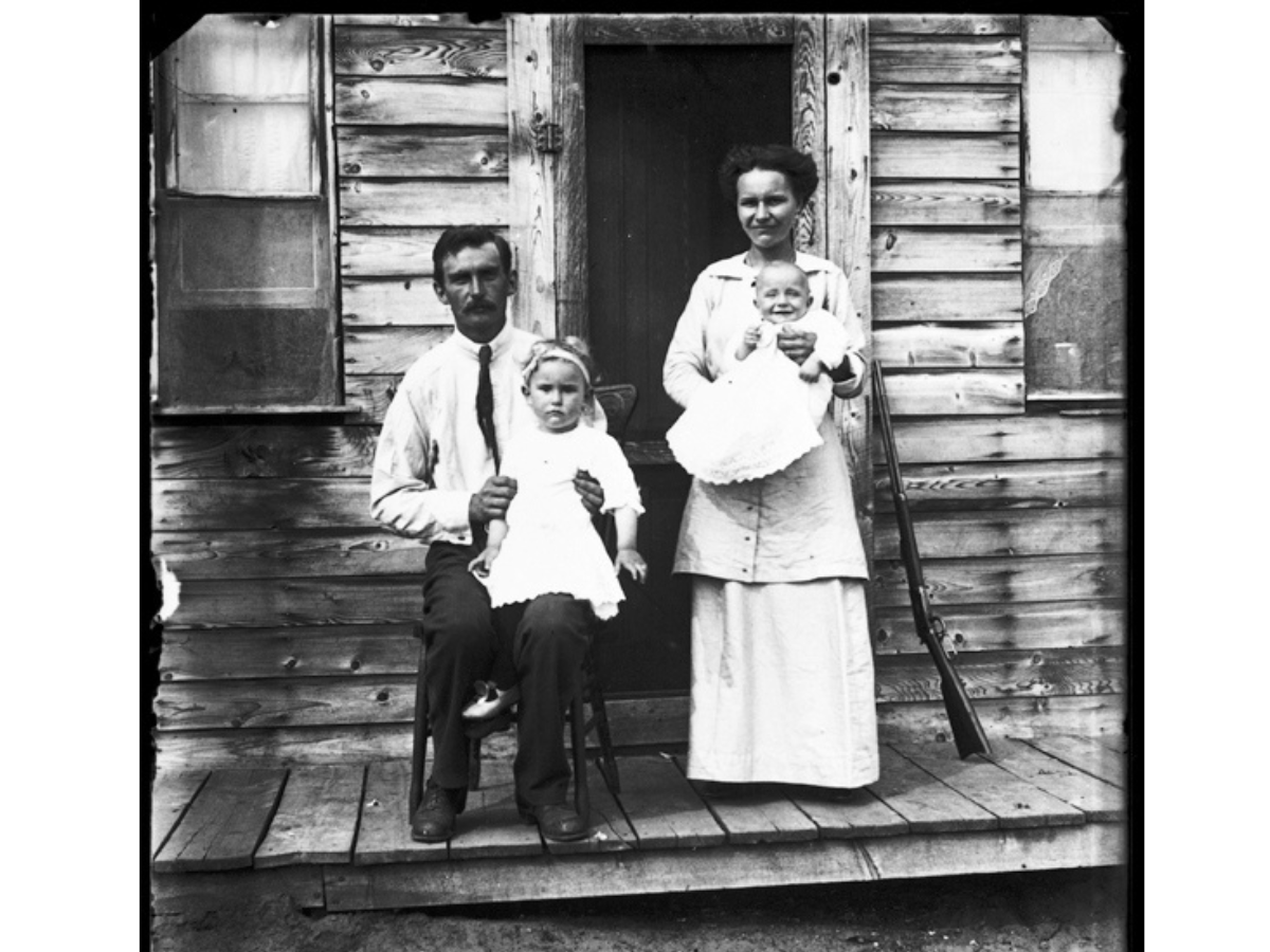 A family on a wooden porch in front the door of a building. A man sits on a chair with a toddler on his knee. Beside him stands a woman holding a smiling baby. A rifle is propped against the doorframe beside them.