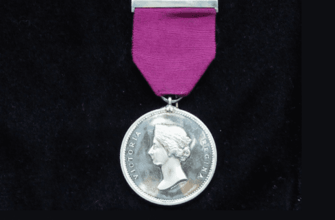 Photograph of circular silver medal with a likeness of young Queen Victoria. Text either side of the likeness reads, “Victoria Regina”. Medal is hung on a piece of purpleish fabric or ribbon.