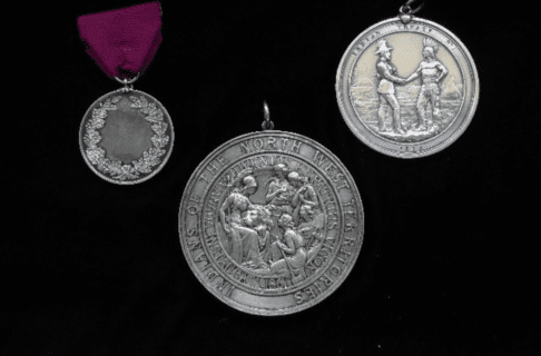 Photograph of three silver medals. L-R: 1. The smallest medal; circular and decorated with an engraving of an oak leaf wreath; hung on a purplish piece of ribbon or fabric. 2. The largest medal; highly decorative, portaying a scene with Imperial Britannia, a lion, and Roman maidens; text around the edge of the medal reads, “Indians of the North West Territories / Juventas et Patrius Vigor / Canada Instaurata 1867”. 3. Final medal; the Treaty No. 1 handshake medal; a circular silver medal portraying a representitive of England shaking hands with a First Nations leader; they stand on grassy ground in front of tipis and the rising sun; text around the edge of the medal reads, “Indian Treaty No. 1 / 1873”.