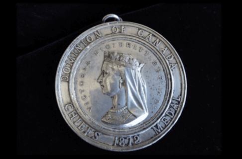 Photograph of a circular, silver medal with a likeness of Queen Victoria. Around the likeness text reads, “Victoria D : G: Britt : Reg : F : D”. Text around the edge of the medal reads, “Dominion of Canada / Chief’s Medal / 1872”