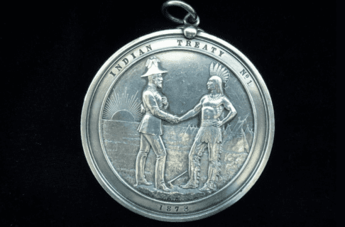 Photograph of a Treaty Number 1 handshake medal. A circular silver medal portraying a representitive of England shaking hands with a First Nations leader. They stand on grassy ground in front of tipis and the rising sun. Text around the edge of the medal reads, “Indian Treaty No. 1 / 1873”.