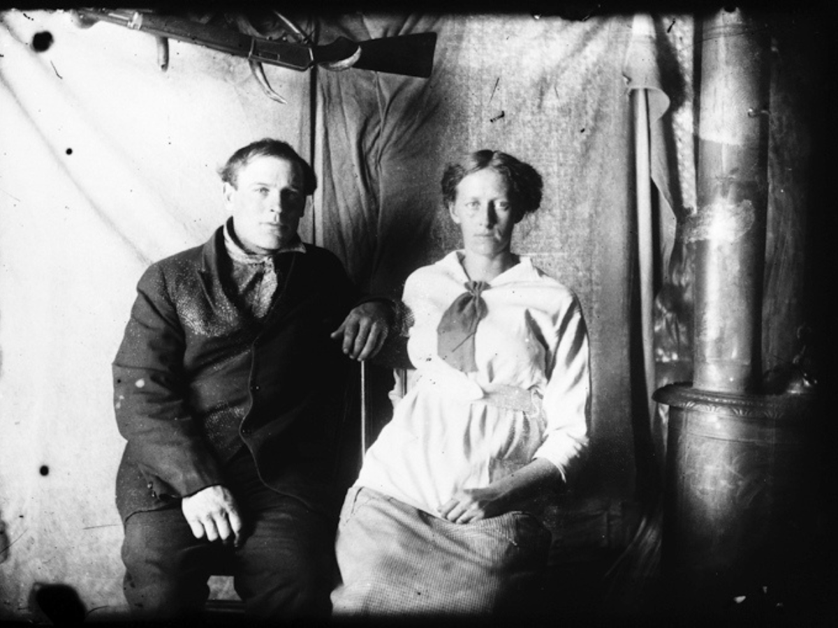 A man and a woman sitting side by side in front of a make-shift hanign backdrop. The man is bundled up, wearing a dark jacket and neck kerchief. The woman is wearing a light-coloured top and apron with a lightly-patterned skirt and a neck kerchief. Both are looking directly into the camera with serious expressions.