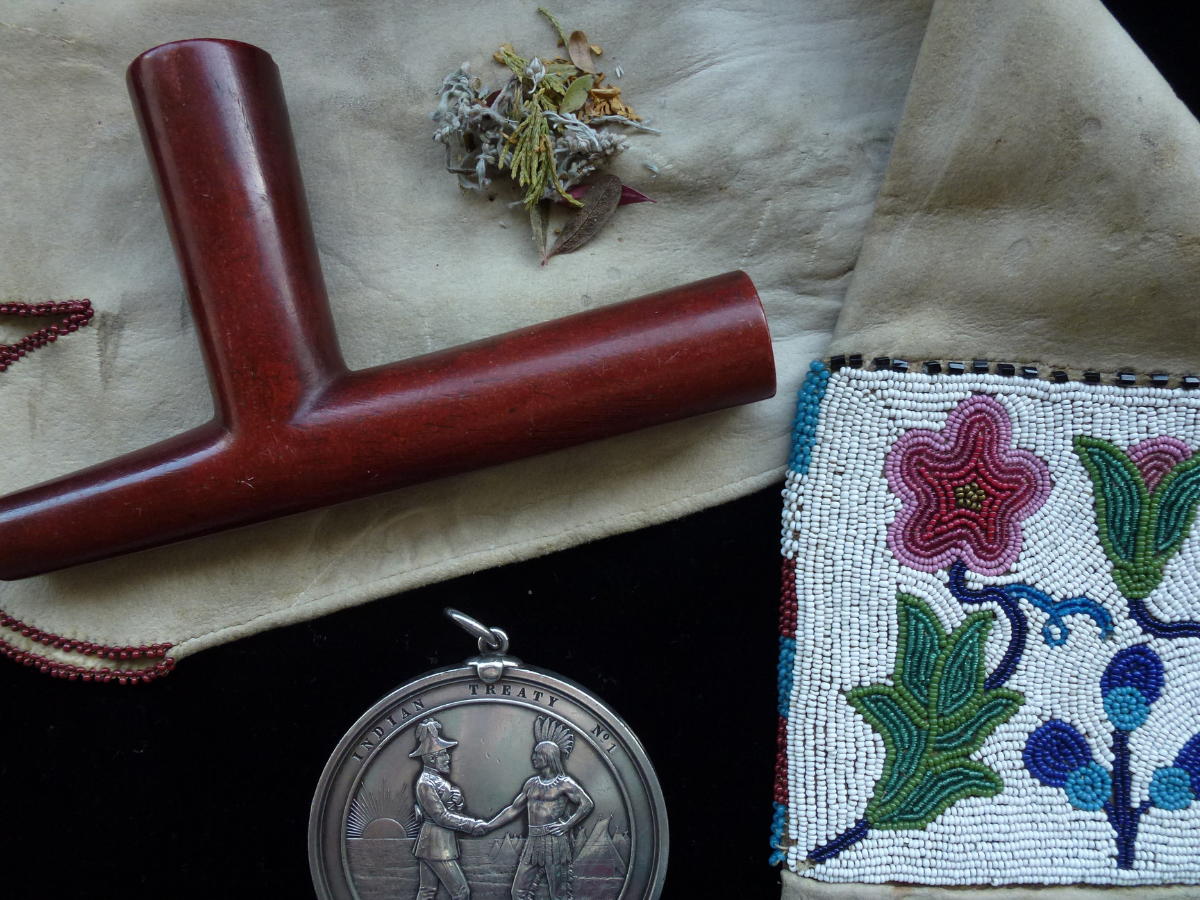 Close-up view of some of the artifacts featured in the “We Are All Treaty People” Exhibit: a wooden pipe and some tabacco laid on a pipe bag with beaded detailing. A Treaty No. 1 handshake medal.