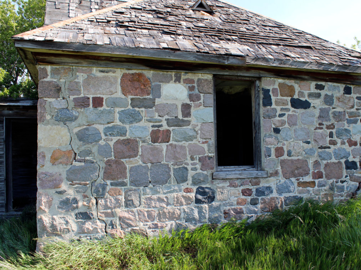 Photograph of the exterior wall of an abandoned stone house with an open window frame and a worn shingled roof.