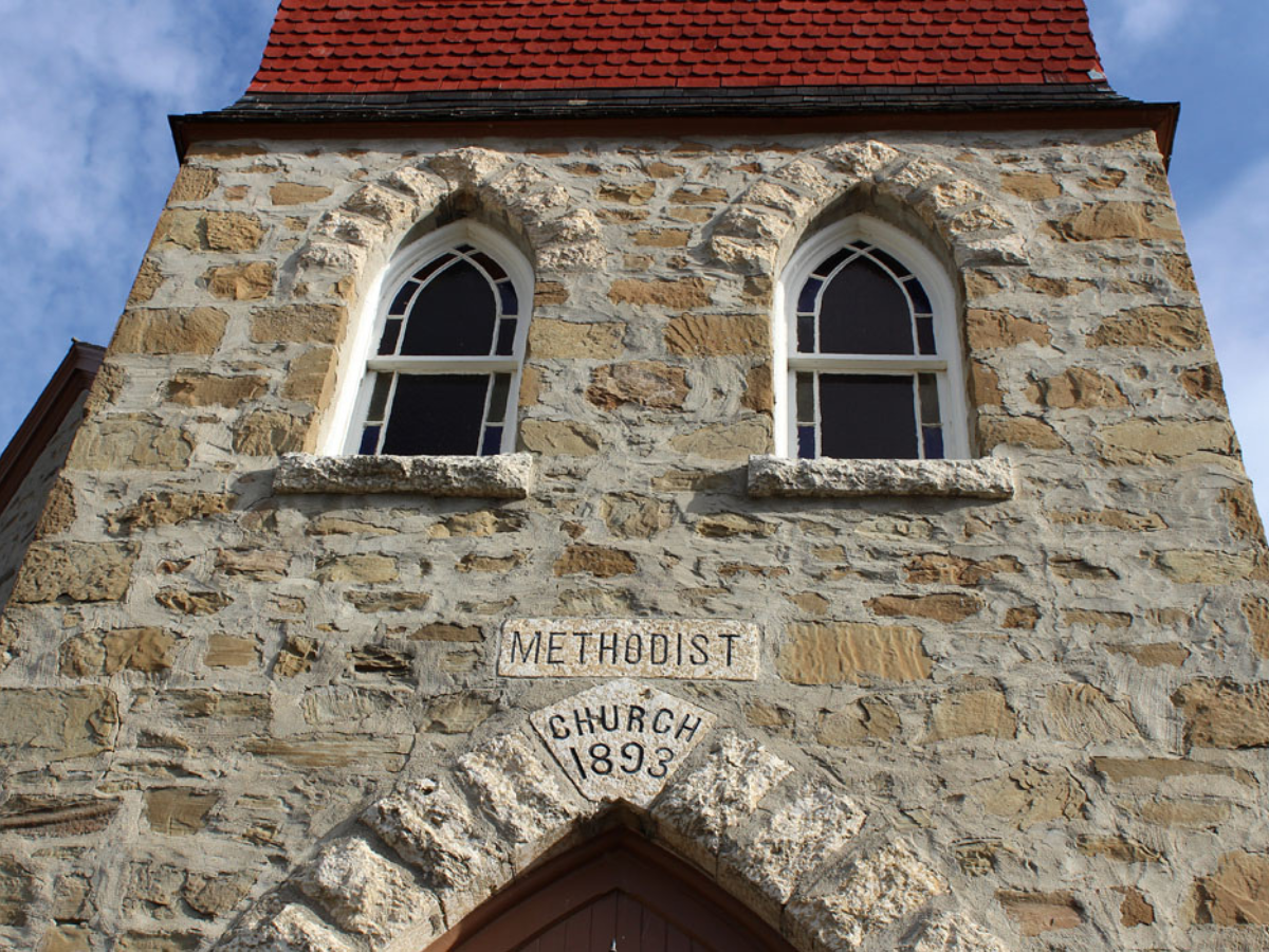 Looking up at the wall of a fieldstone building with two windows side by side. At the bottom of the frame, above the doorway, a datestone reads “Methodist Church / 1893”.