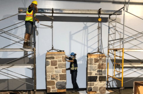 Two “pillars” of frame with fieldstone attached to the exterior being moved into place in the new Prairies Gallery with a hoist and girder system.