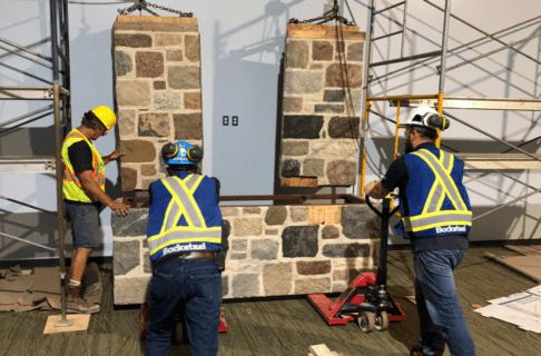 Three individuals maneuver the rectanulgar base of the metal frame and fieldstone wall under the two side pillars, which are hanging in place on a joist and girder system, using two pallet jacks.