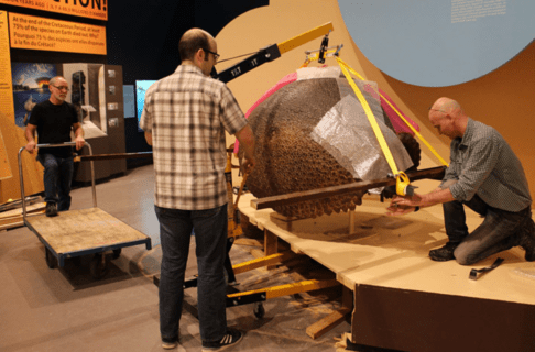 Three individuals work together to move a large Glyptodont carapace onto a wheeled cart from a platform using an engine hoist lift.