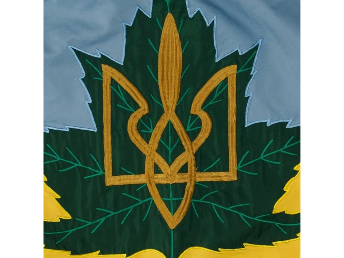 Close-up image of the Ukrainian Canadian Veteran’s flag. The top half of the flag is light blue, and the bottom half is yellow. In the centre is a dark green maple leaf with a gold symbol on it – the Tryzub, or Ukrainian “trident” symbol.