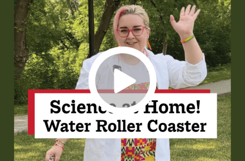 A screenshot of a video, an individual wearing a lab coat waves to the camera while standing outside in front of trees. There's a play button over the screenshot and overlaid text reads, "Science at Home! Water Roller Coaster".