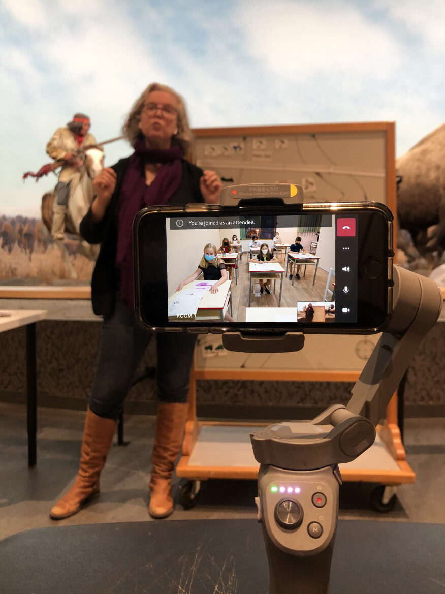 A Museum staff person standing in front of a rolling white board in the Museum Galleries presenting a virtual class to a group of students visible on a smartphone held in frame.
