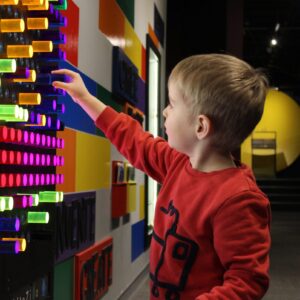 A child placing colourful pegs into the slots of a giant Lite Bright set embedded in a wall of the Science Gallery.
