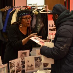 A Black woman wearing a flat-topped hat with a blue and purple woven design around the bottom, stands behind a booth with vintage photographs of Black individuals. She is in discussion with a visitor at the table.