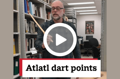A screenshot of a video, an individual standing in a storage area, holding up a section of an atlatl. There's a play button over the screenshot and overlaid text reads, "Atlatl dart points".