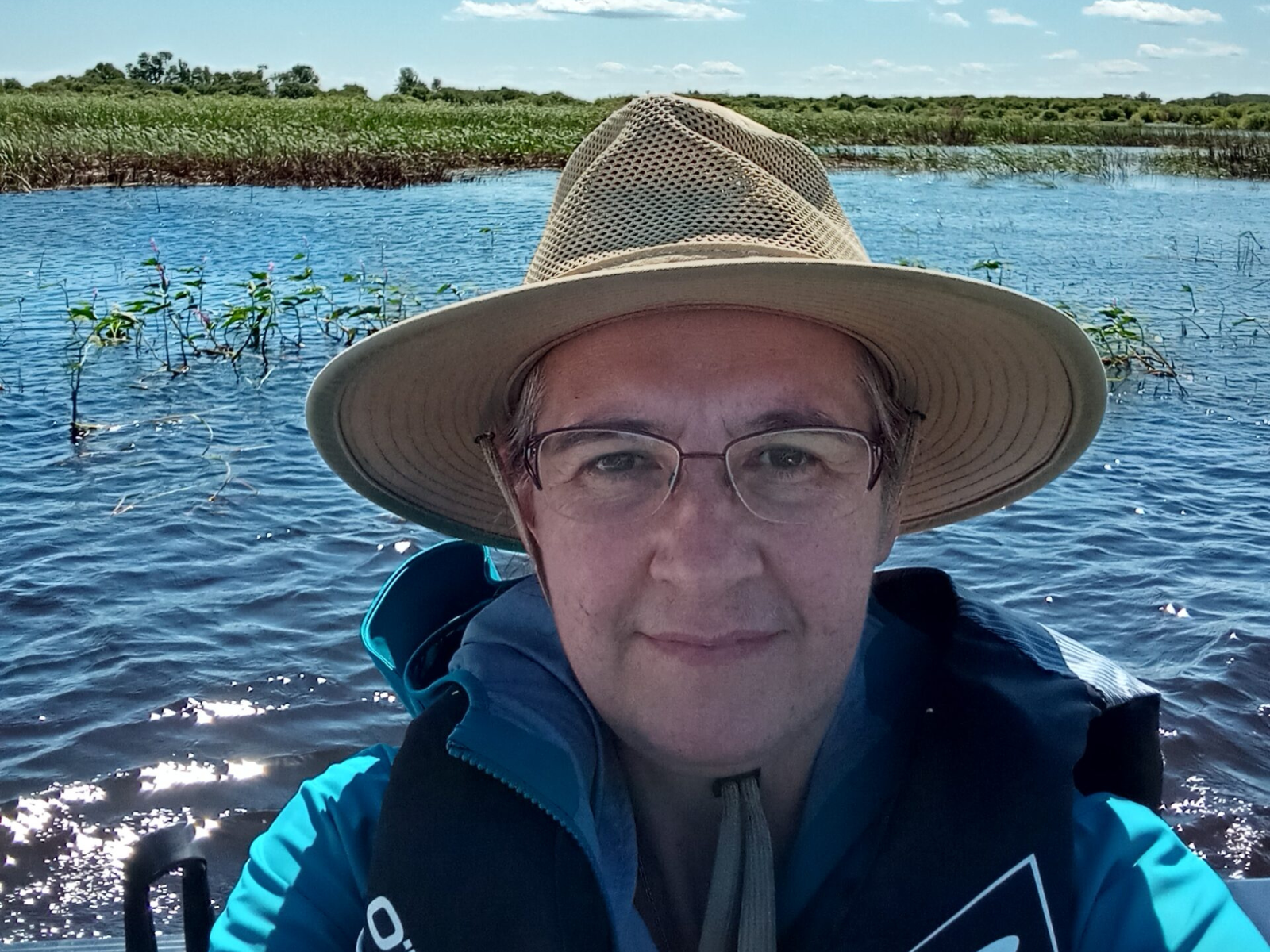 A selfie taken by Dr. Diana Bizecki Robson showing her sitting on a boat with a body of water and shoreline in the background. She is wearing a life jacket, a wide-brimmed sun hat, and glasses.