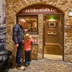 An older adult and a child look through the window of an immersive exhibit set up as an 1920s taxidermist.