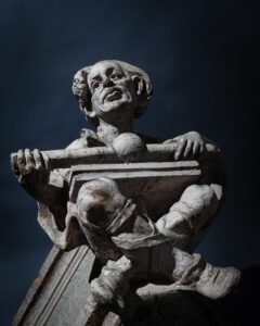 A stone grotesque seated cross-legged with printing materials on his lap.