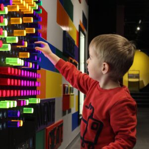 A child placing colourful pegs into the slots of a giant Lite Bright set embedded in a wall.