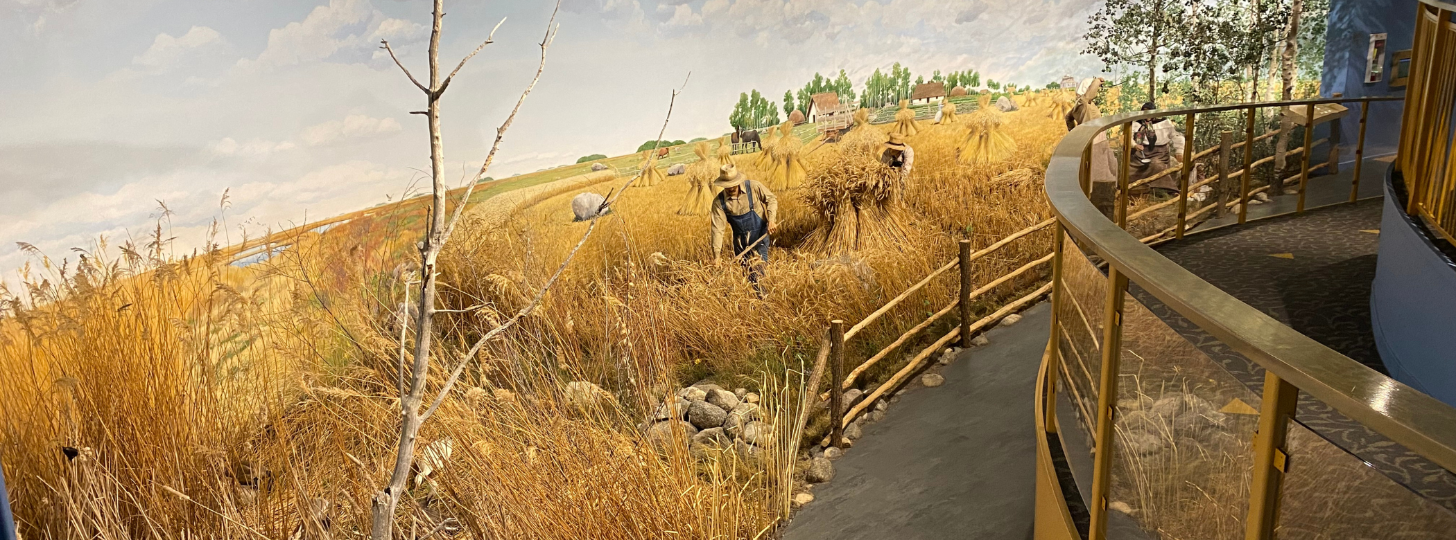 Diorama depicting a man wearing blue coveralls and a white button up shirt, a straw hat on his head holding a scythe in his hand. The man is using the scythe to cut down a field of wheat stalks. Boulders are in the forefront and green prairie grasses are scattered about.