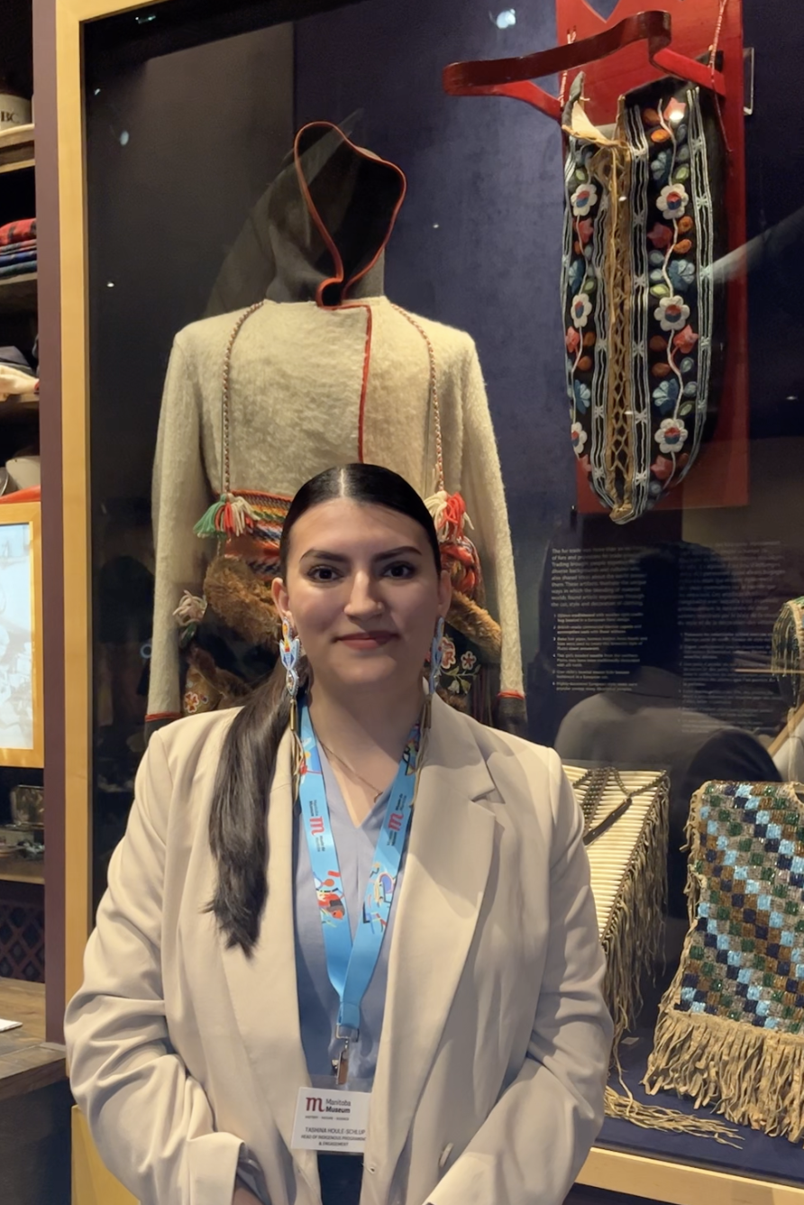 A Museum staff person standing in front of a display case containgin a number of Indigenous artifacts, including a cradleboard.