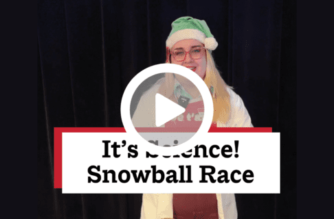 A screenshot of a video, an individual wearing a white lab coat and green Santa hat. There's a play button over the screenshot and overlaid text reads, "It's Science! Snowball Race".