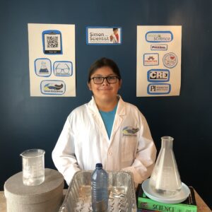 A smiling youth wearing a white lab coat standing in front of a table with various science experiment implements laid out, like beakers.