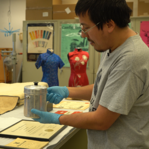 An individual standing in a collections lab wearing blue gloves holds up a small tin lunch container. Two Chinese dresses, red and blue, are on dress forms in the background.