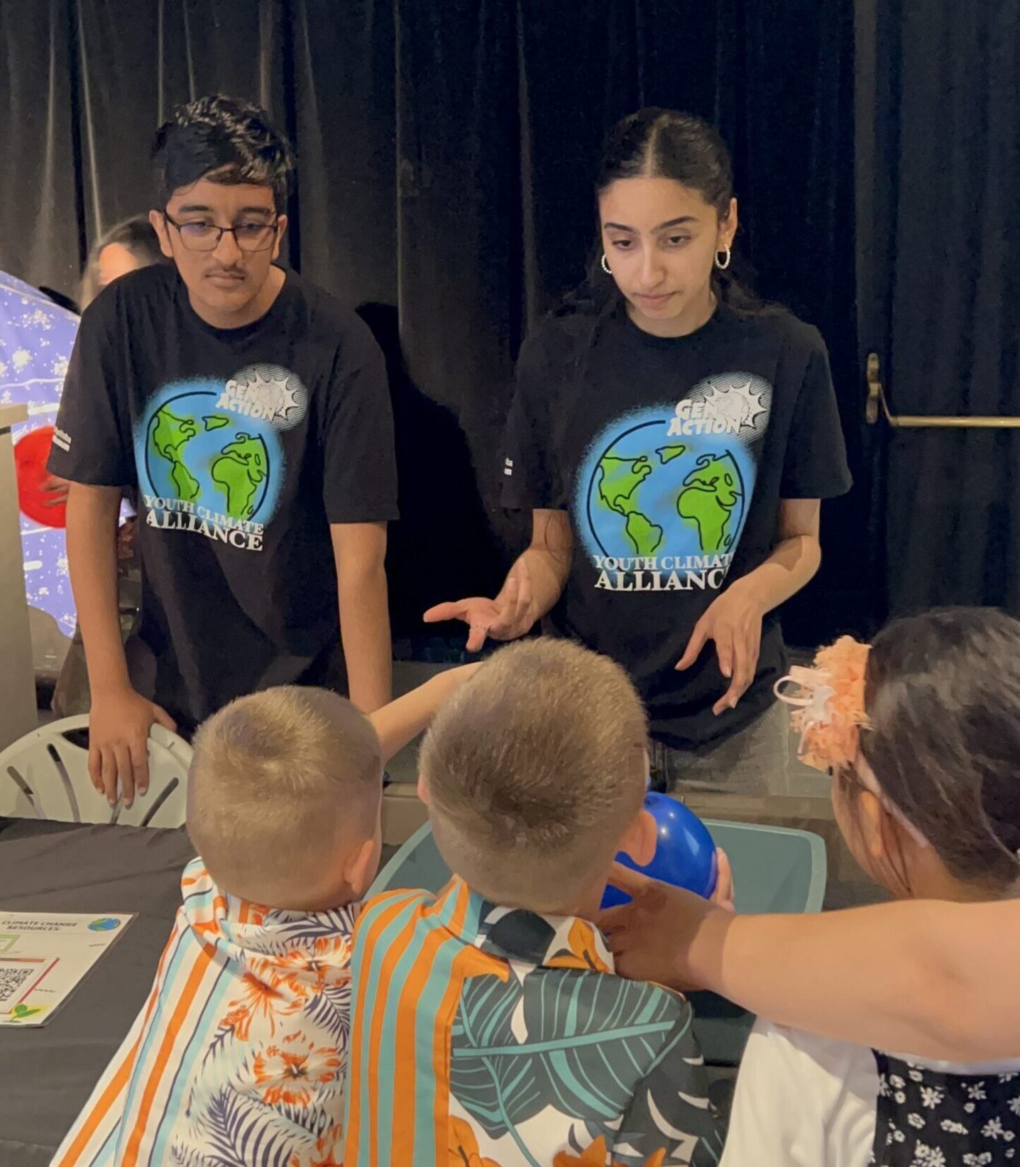 Two youth wearing Youth Climate Alliance t-shirts engage with three young visitors at a pop-up exhibit.