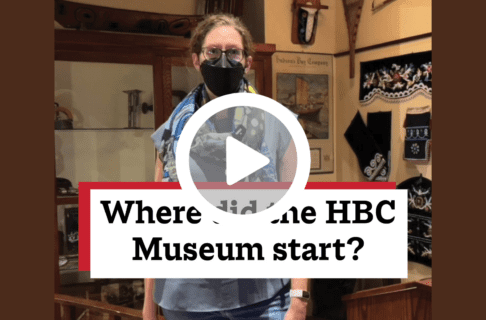 A screenshot of a video, an individual standing in the HBC Museum Collection Gallery. There's a play button over the screenshot and overlaid text reads, "When did the HBC Museum start?".