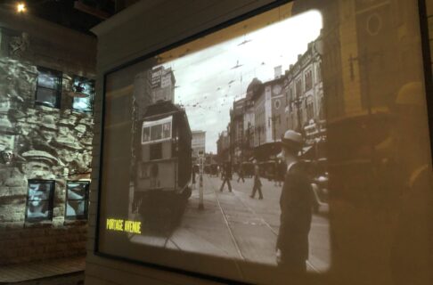 Black and white video footage of traffic on a street in the early 1900s projected onto a faux building exterior. The footage is labelled "Portage Avenue".