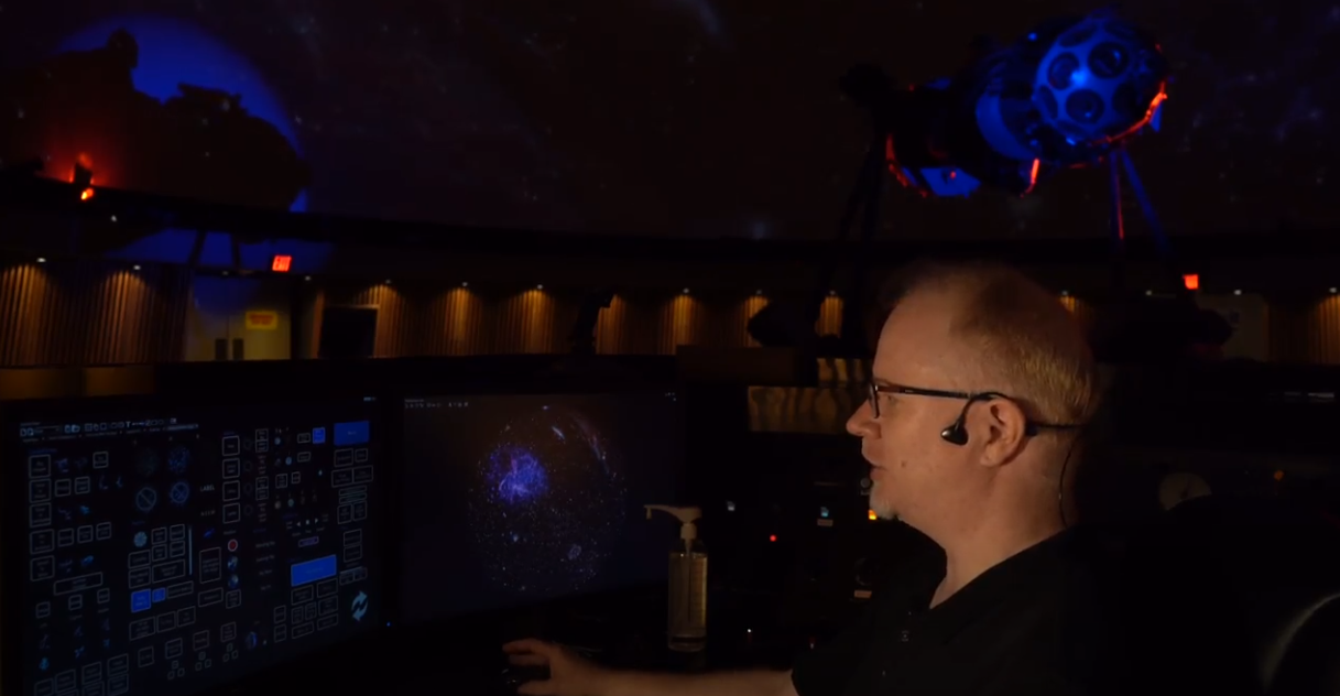 An individual wearing a headset sitting at a desk with two computer monitors under a darkened planetarium dome.