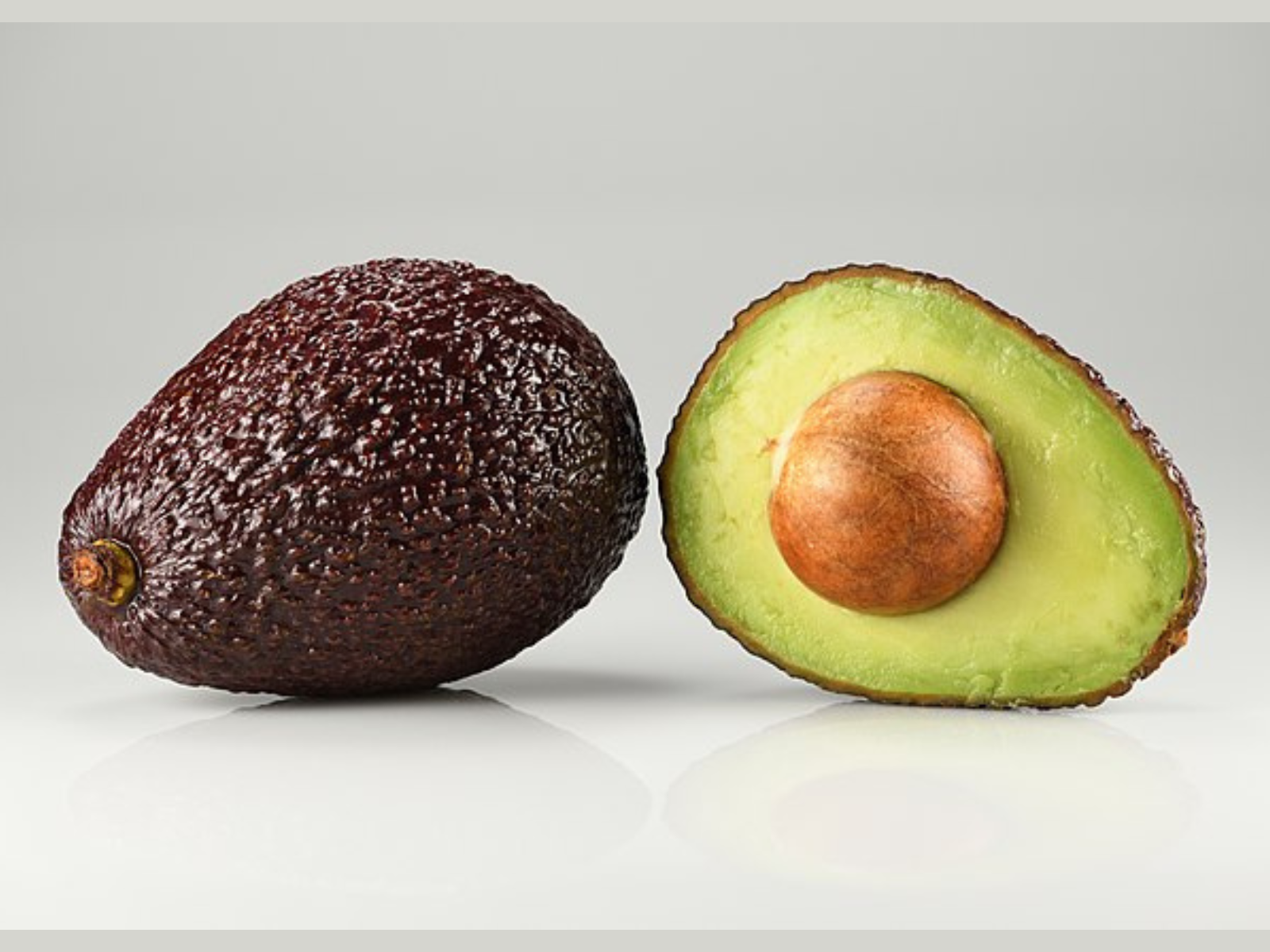 Two avocados on a grey background. The avocado on the left is whole, with purple-brown skin, and the one of the right has been cut in half showing the green flesh and round brown seed.