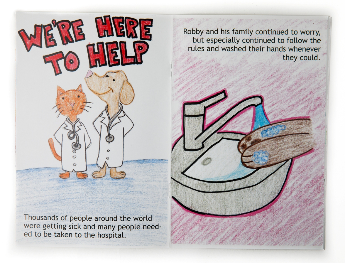 Pages from a hand-drawn book. On the right the page has a drawing of a cat and a dog wearing white lab coats and stethoscopes. In large letters above their heads it says, "We're here to help". Printed text at the bottom reads, "Thousands of people around the world were getting sick and many people needed to be taken to the hospital." The page on the left show paws washing with soap and water in a sink. Printed text along the top reads, "Robby and his family continued to worry, but especially continued to follow the rules and washed their hands whenever they could."