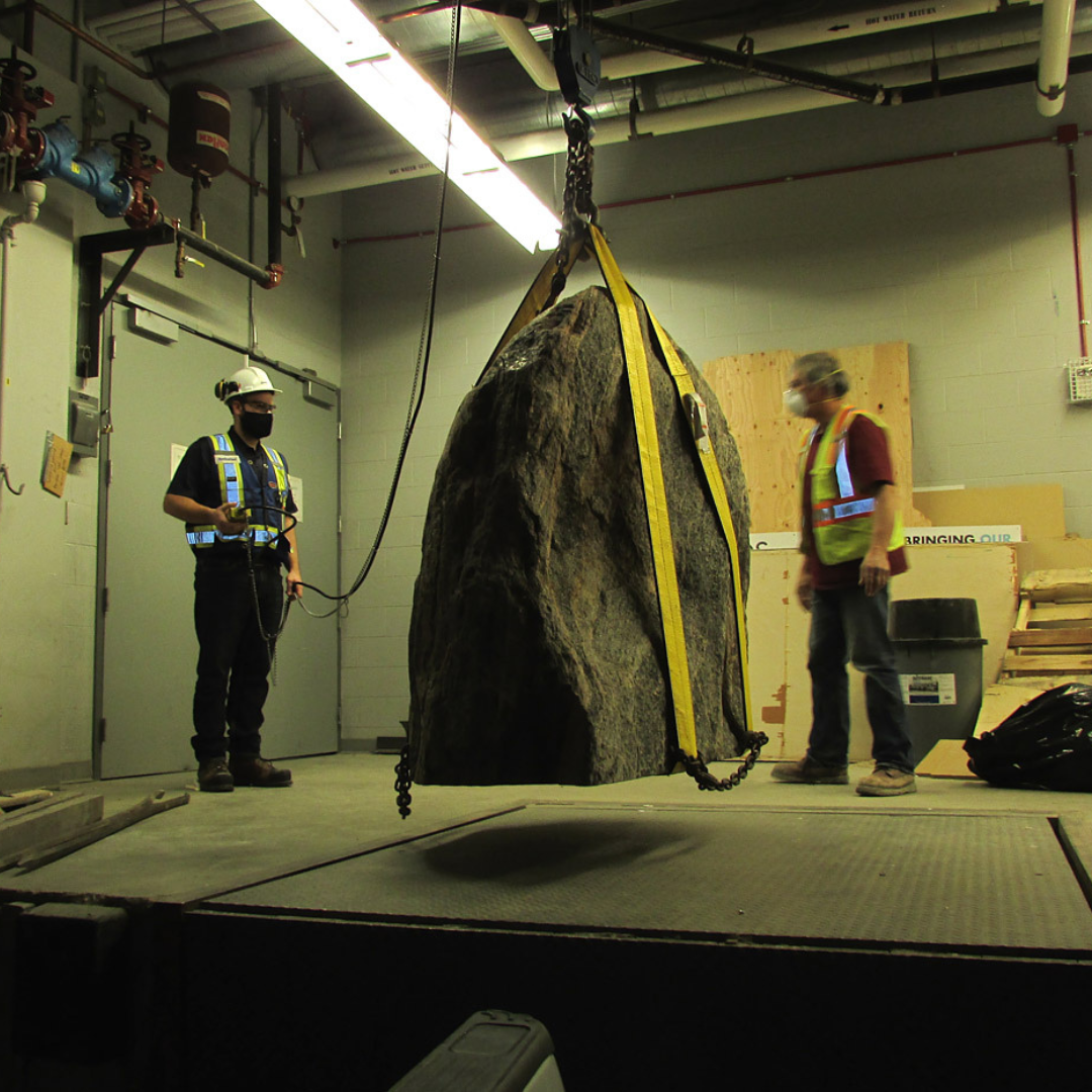Two individuals wearing high-vis vests observe as a large boulder is lifted over a loading dock platform with a dock hoist operated by one of them.