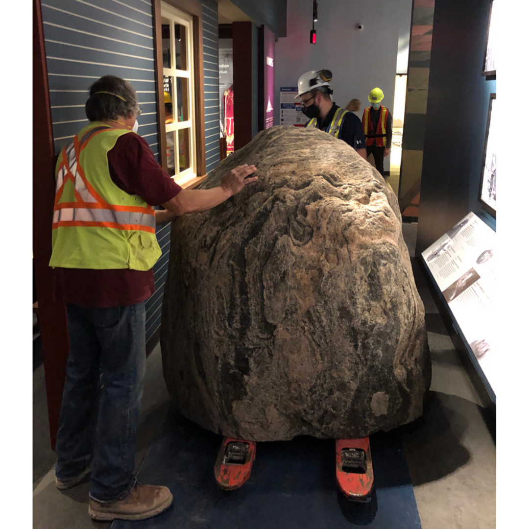 Three individuals in high-vis vests guide a large boulder secured on a pallet jack through a narrow space in the Winnipeg Gallery.