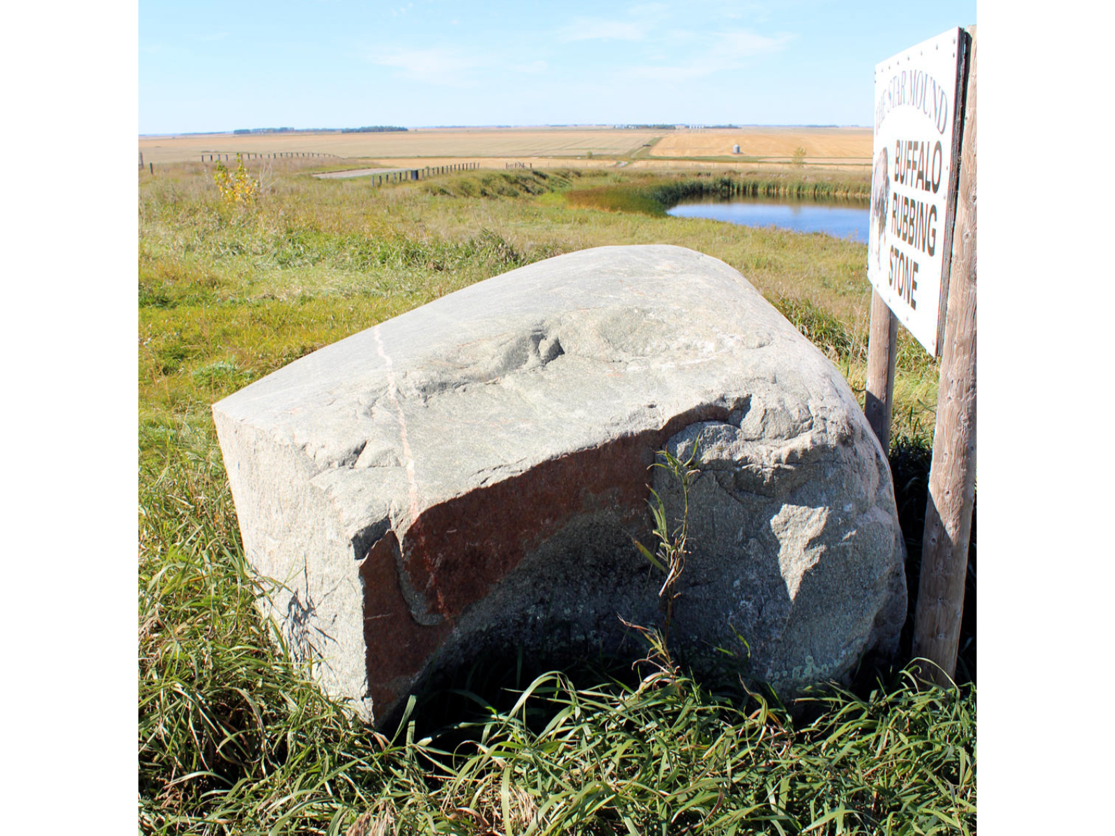 A large grey boulder in the grass next to a sign angled away from the camera that cannot be read. The top and sides of the boulder are slightly rounded.