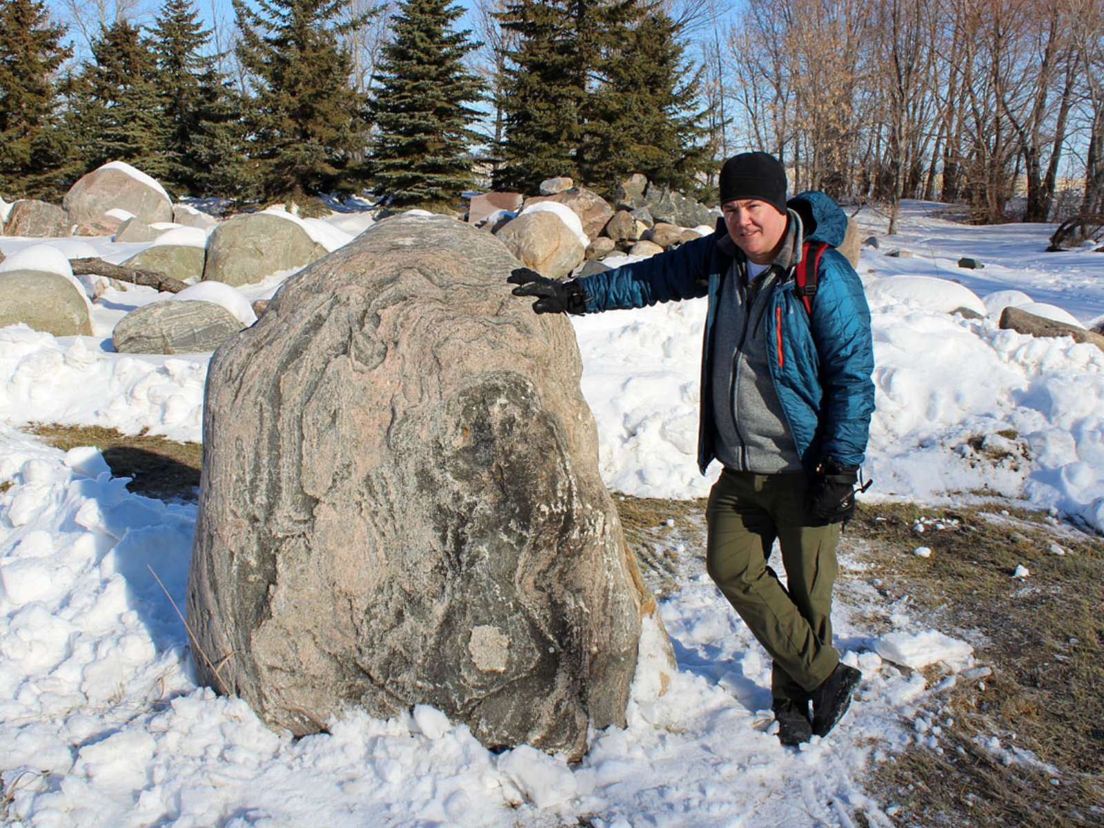 A person wearing winter gear smiles at the camera and leans against a large boulder that comes as high as their shoulder.