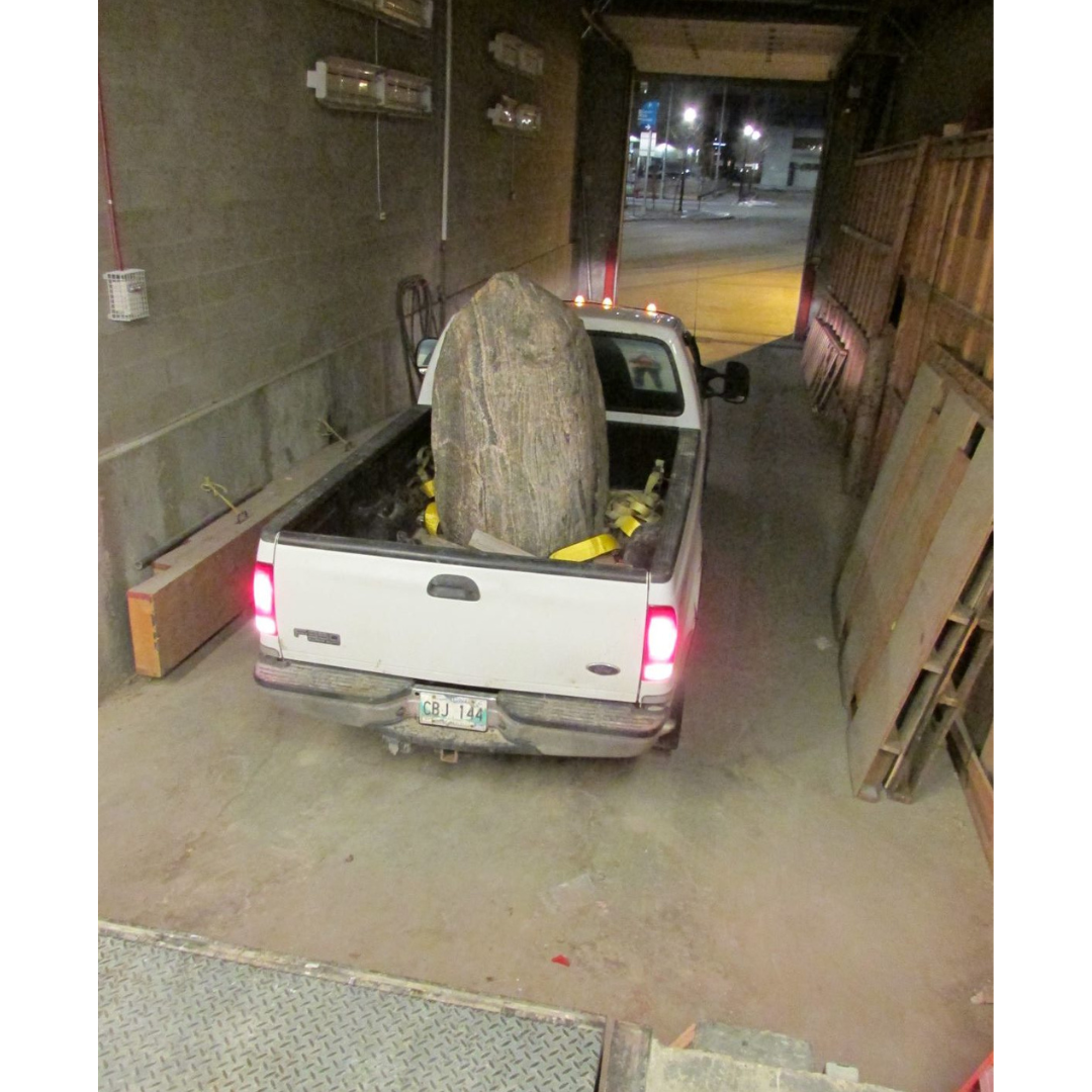 A white pick-up truck backed into an enclosed loading zone with a large boulder in the back of the truck.