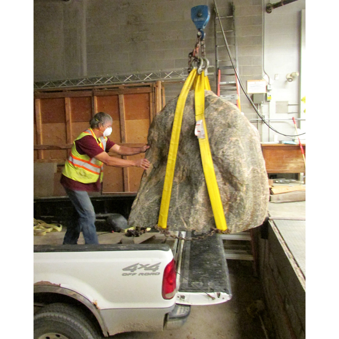 A large boulder being lifted with a dock hoist from the back of a white pick-up truck. An individual wearing a high-vis vest guides the boulder off the truck.