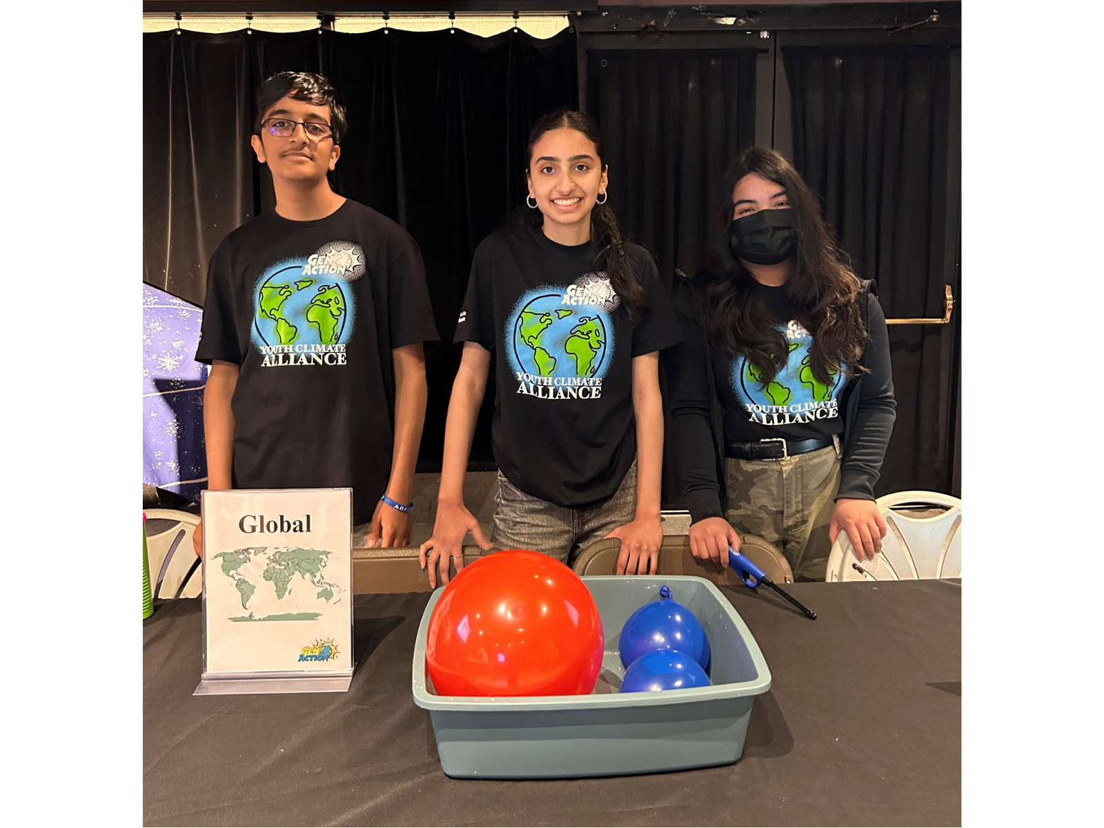 Three youth wearing Youth Climate Alliance t-shirts stand behind a pop-up exhibit table with a container of water and two balloons floating in front of them.