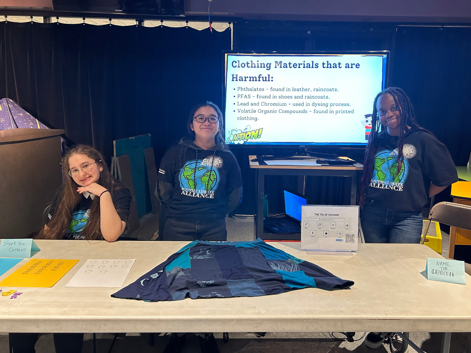 Three youth wearing Youth Climate Alliance t-shirts stand behind a pop-up exhibit table with a shirt laid out in front of them. On a screen behind them text reads, "Clothing Materials that are Harmful:"
