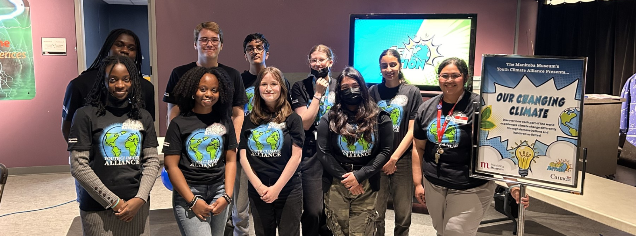 A group of nine youth and a Museum staff member smiling together. All are wearing matching t-shirts with an illustrated globe and the words “GenAction! / Youth Climate / Alliance”. On the right side of the group is a sign reading, “Our Changing Climate”.
