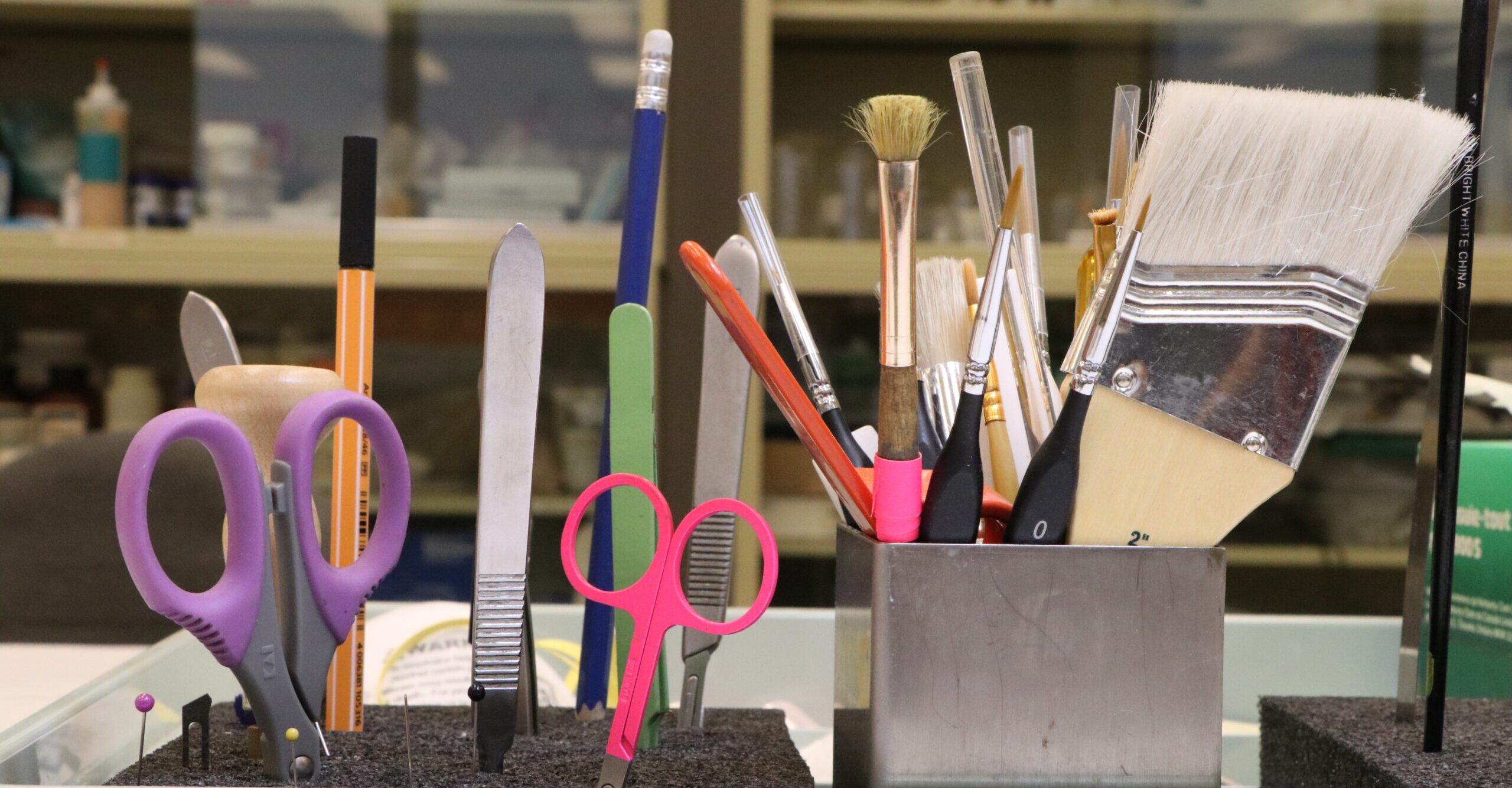 Scissors, scalpel blades, and various sized paint brushes sitting in stainless steel holder.