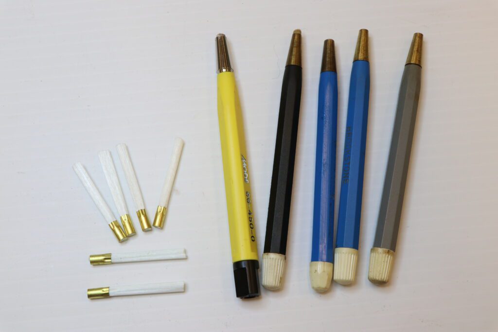 Five glass bristle brush dispensers, from left to right the handles are yellow, black, blue, blue and grey with a white twist bottom used to make the brush larger. On the left of the image are five glass bristle brush replacements with a gold coloured ferrule crimping the bristles together. 