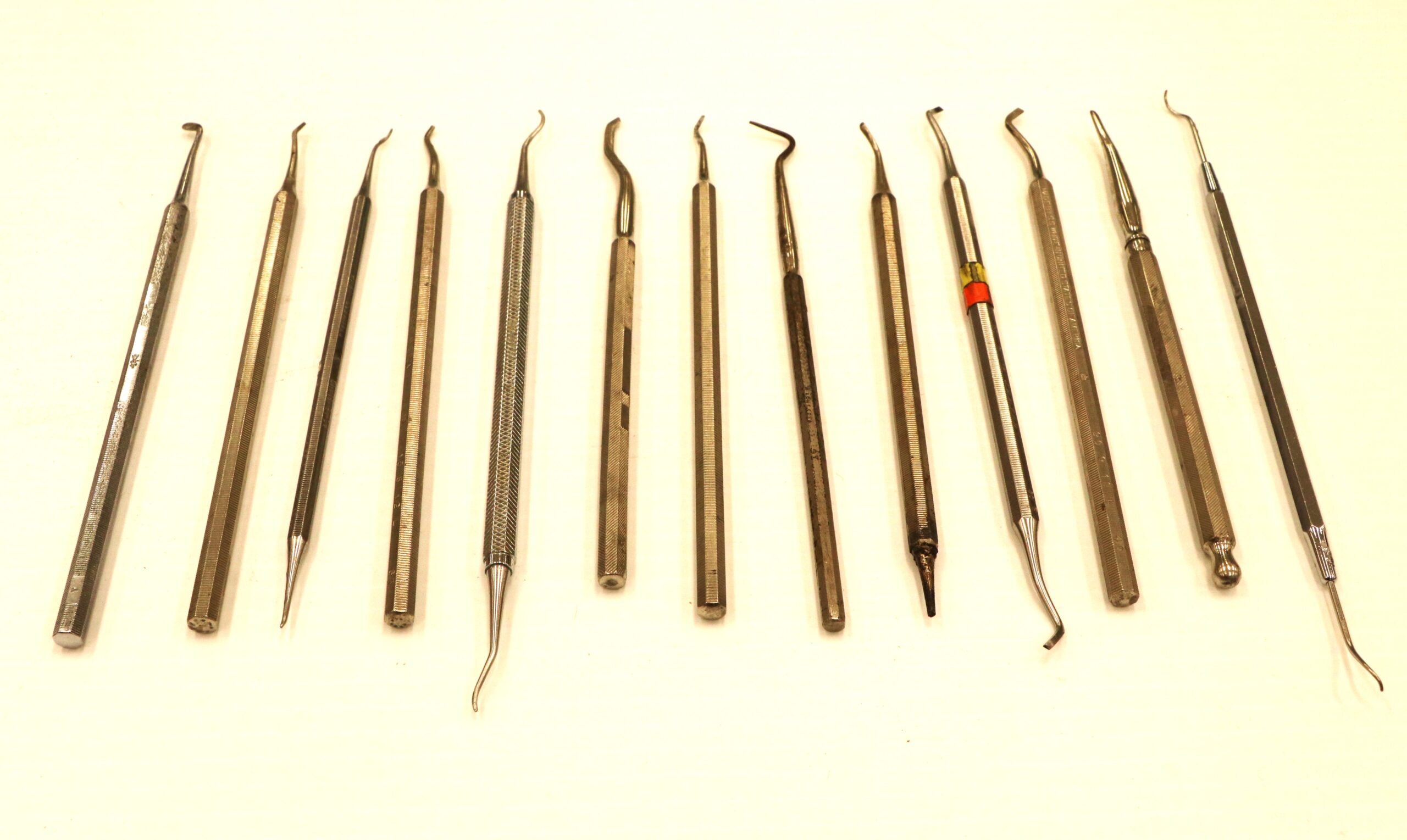 Image of thirteen dental tools laid out evenly in a row. Tools are composed of stainless steel and have various tips on both ends