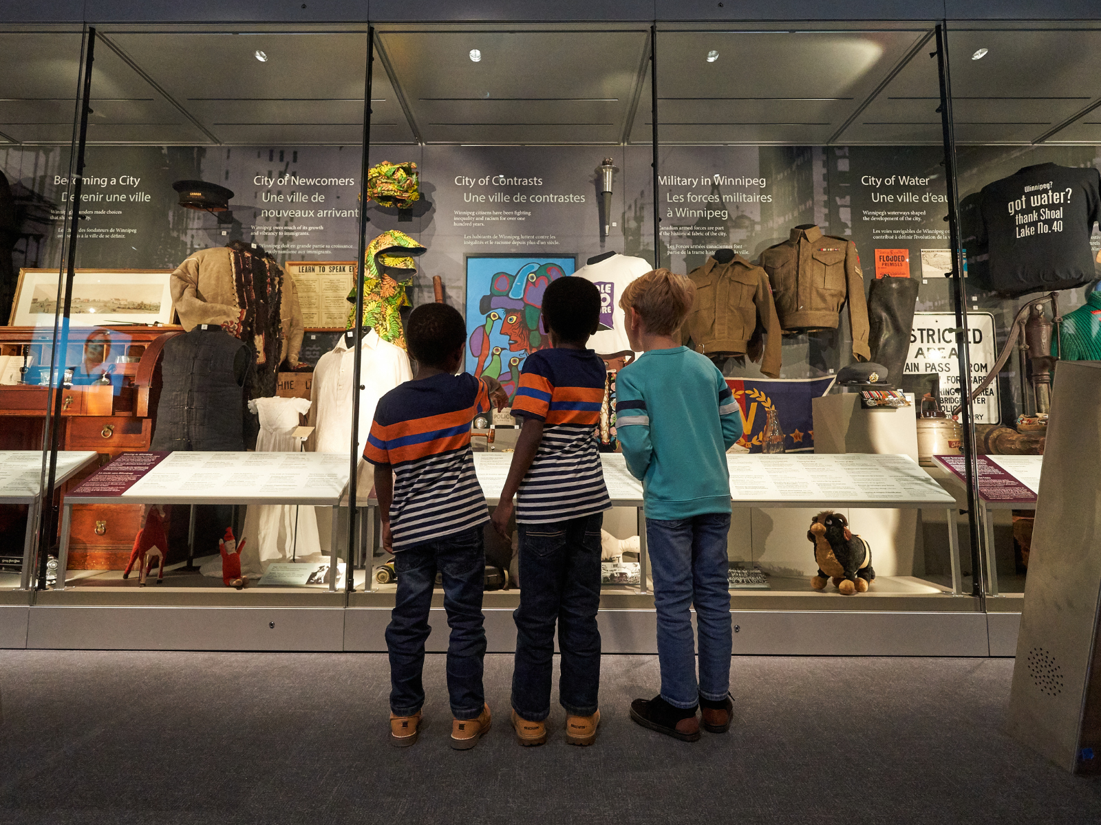 From behind, three children standing together looking into a wall of display cases filled with artifacts.
