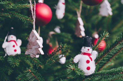 Close-up on a snowman ornament on a Christmas tree. Other ornaments are out of focus in the background including other snowmen, wooden trees, and red baubles.